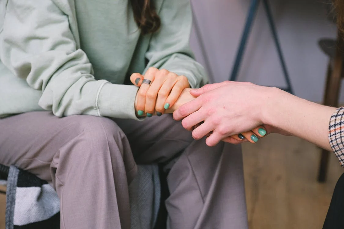 Close-up of two people's hands clasped together in a supportive gesture, one wearing a green sweater and the other with teal nail polish. Close-up of two people's hands clasped together in a supportive gesture, one wearing a green sweater and the other with teal nail polish.