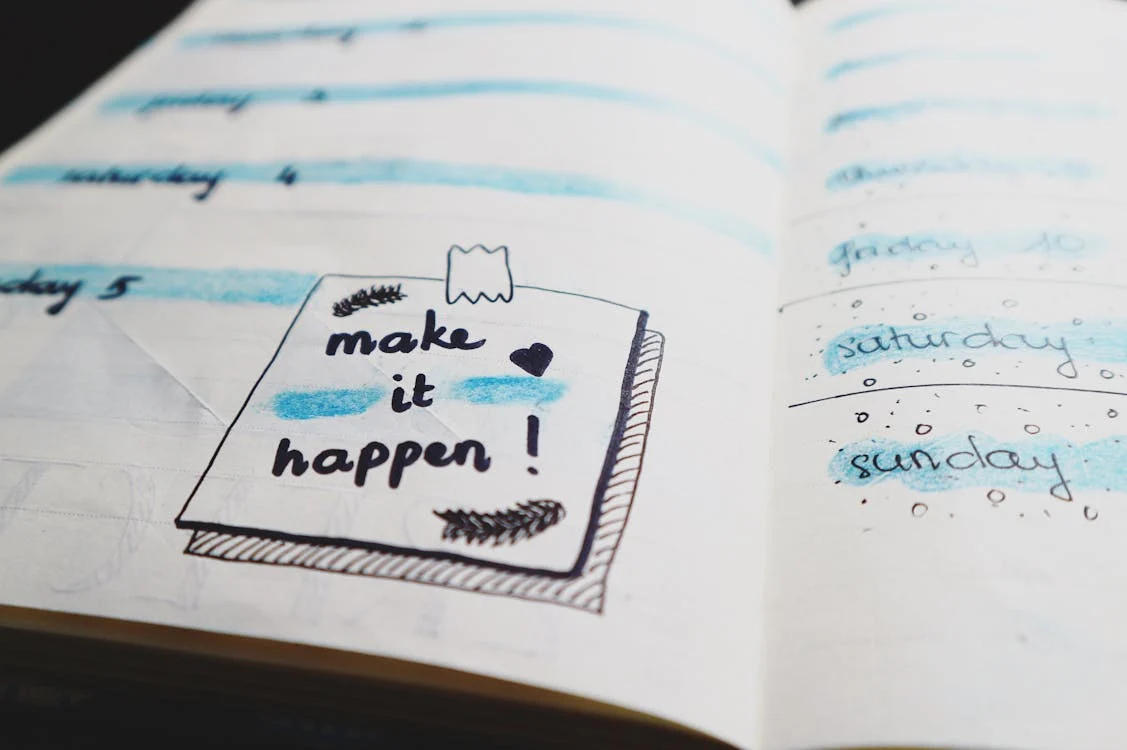 A planner page with "make it happen!" written on a sticky note, surrounded by days of the week and blue highlights. A planner page with "make it happen!" written on a sticky note, surrounded by days of the week and blue highlights.