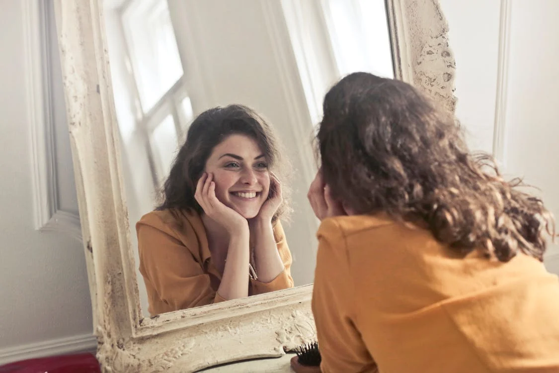 A smiling young woman with dark hair looking at herself in an ornate mirror, wearing a yellow top. A smiling young woman with dark hair looking at herself in an ornate mirror, wearing a yellow top.