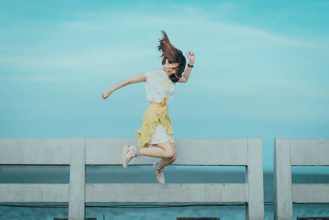 A woman in a white top and yellow skirt jumping joyfully on a concrete barrier against a bright blue sky. A woman in a white top and yellow skirt jumping joyfully on a concrete barrier against a bright blue sky.