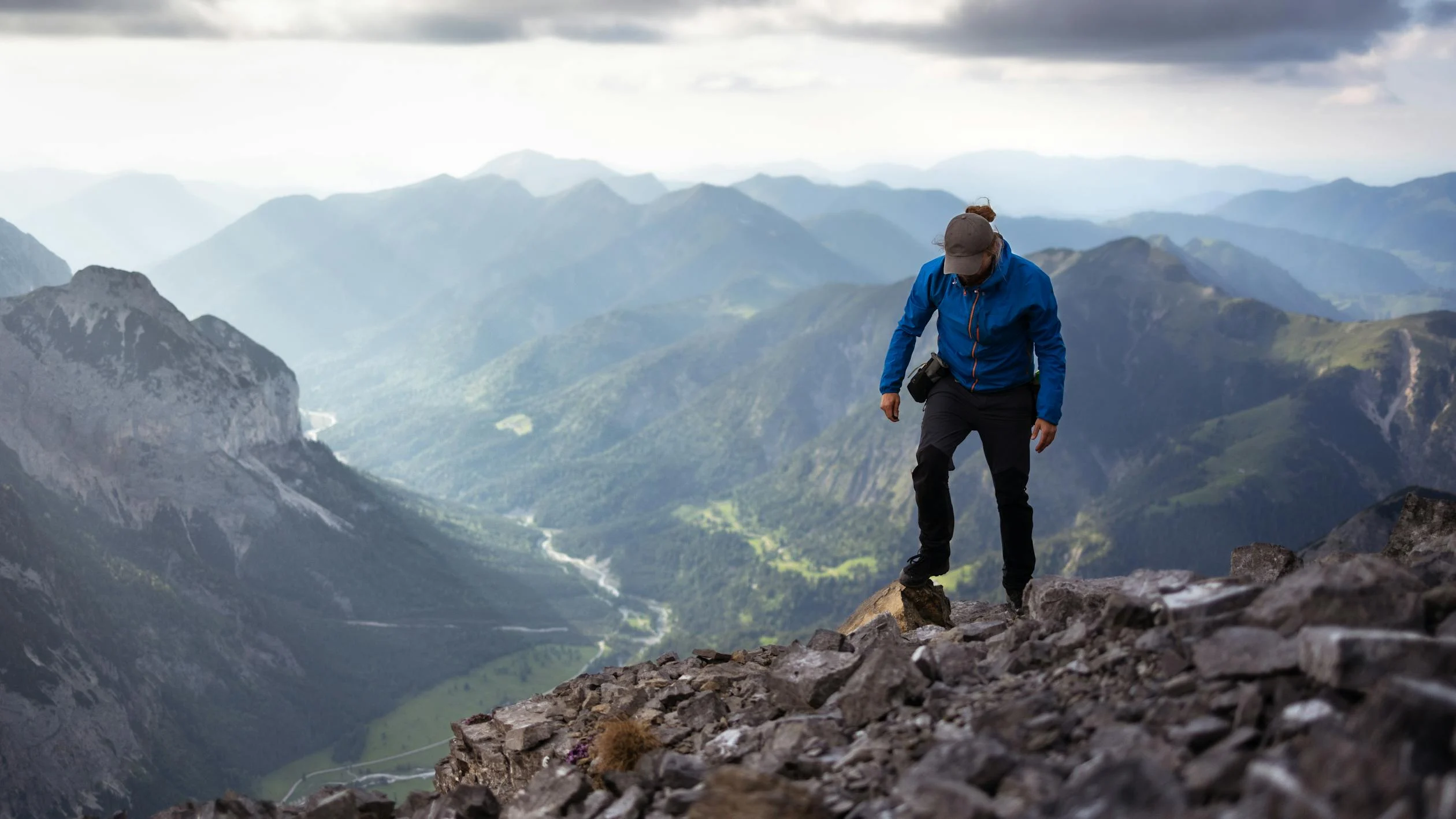 Hiker in blue jacket standing on rocky mountain summit, looking down at a breathtaking panorama of mountain ranges and valleys stretching into the distance under a cloudy sky. Hiker in blue jacket standing on rocky mountain summit, looking down at a breathtaking panorama of mountain ranges and valleys stretching into the distance under a cloudy sky.