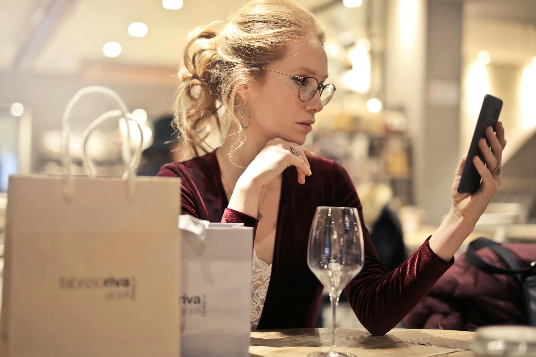 Woman with glasses and blonde hair in a burgundy velvet top intently looking at her smartphone while sitting at a restaurant table with a wine glass and shopping bags nearby. Woman with glasses and blonde hair in a burgundy velvet top intently looking at her smartphone while sitting at a restaurant table with a wine glass and shopping bags nearby.