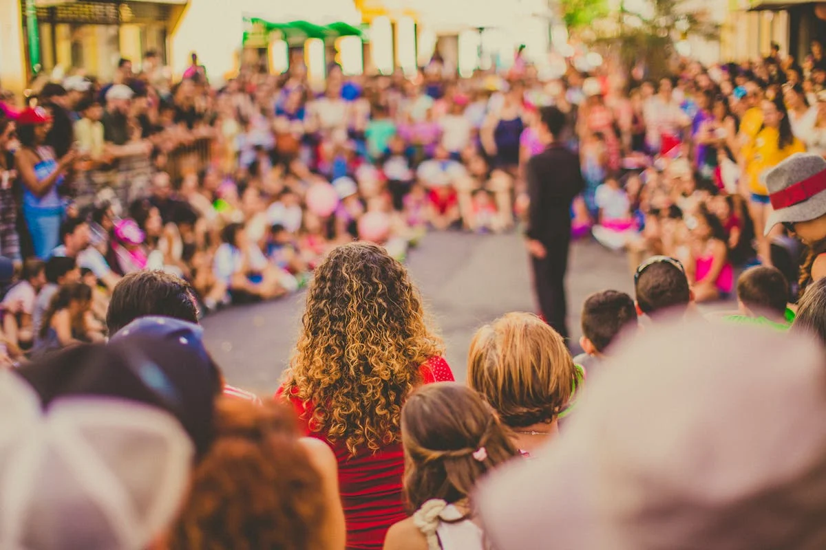 Crowd watching a performance from behind, showing backs of heads with a person in a red striped shirt with curly hair in focus, and a blurred audience in the background with warm lighting and festive atmosphere. Crowd watching a performance from behind, showing backs of heads with a person in a red striped shirt with curly hair in focus, and a blurred audience in the background with warm lighting and festive atmosphere.